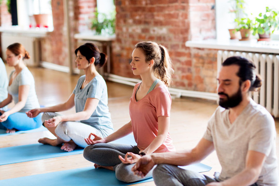 3 women and 1 man sit cross legged on yoga mats with their eyes closed in a well lit room.