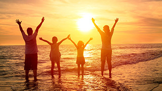 2 adults and 2 children stand in front of an ocean at sunset and hold their arms up in joy.