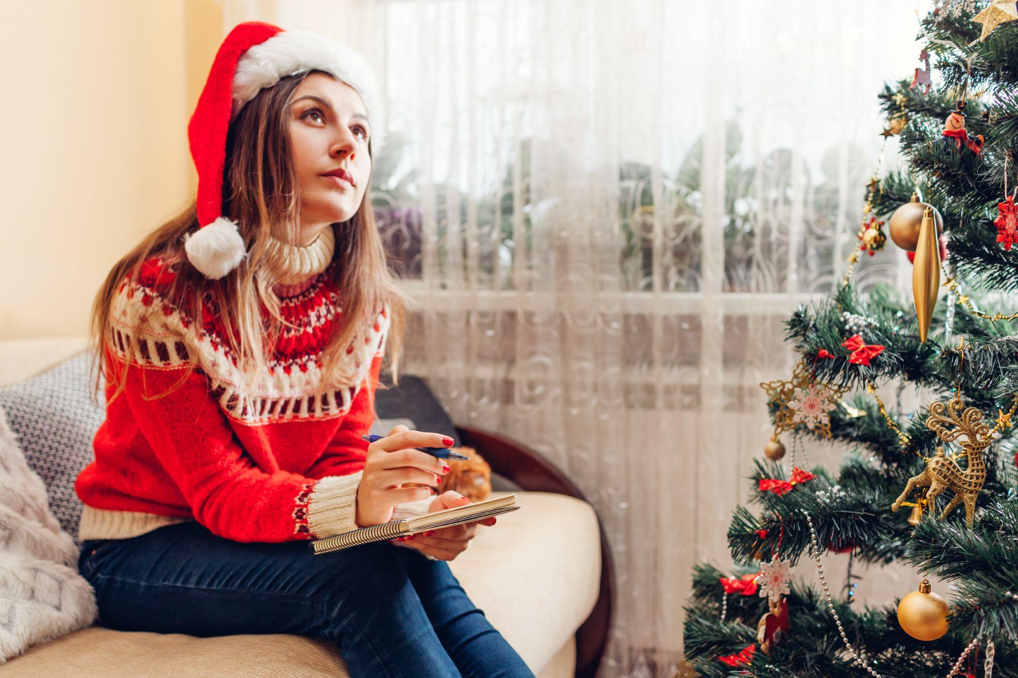 Young woman writing goals for New year in notebook at home sitting by Christmas tree in Santa hat.