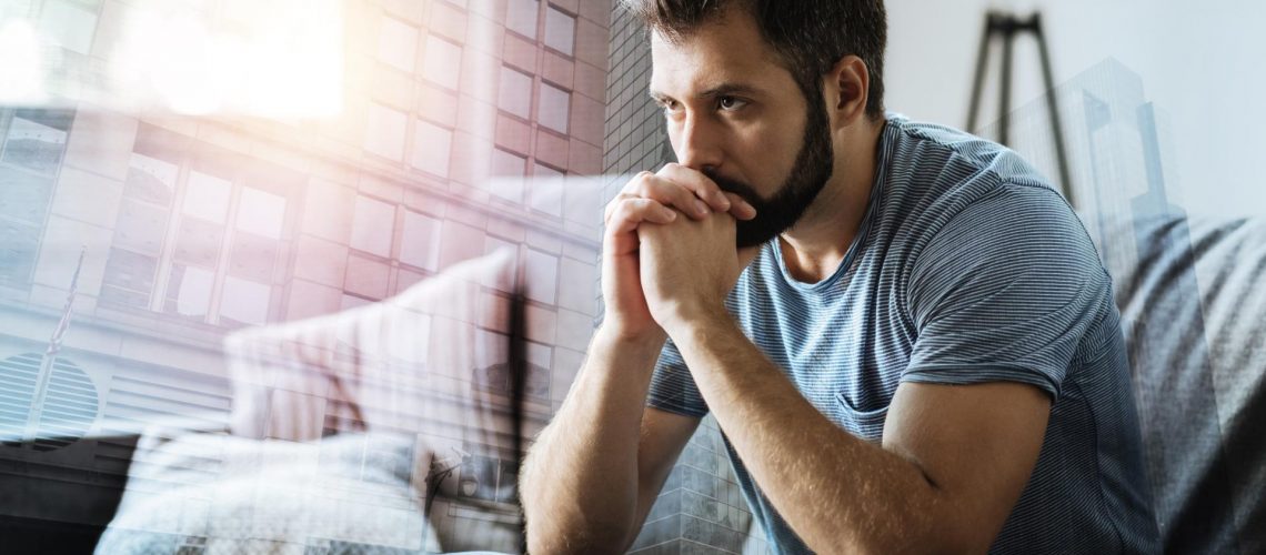 Thoughtful serious man sitting in the empty room holding hands near mouth and thinking.